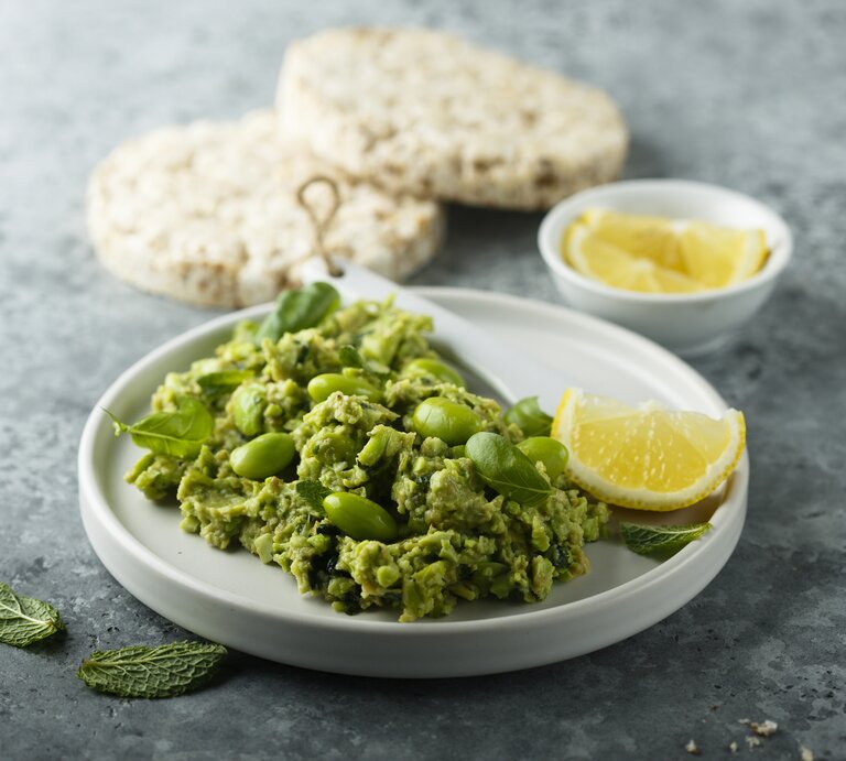 Frontal: eine weiße Schale Edamame-Guacamole steht auf einem grauen Untergrund. Sie ist mit einer Zitronenscheibe garniert. Im Hintergrund liegen Reiswaffeln und noch mehr Zitronenscheiben. | © Getty Images / Mariha-kitchen