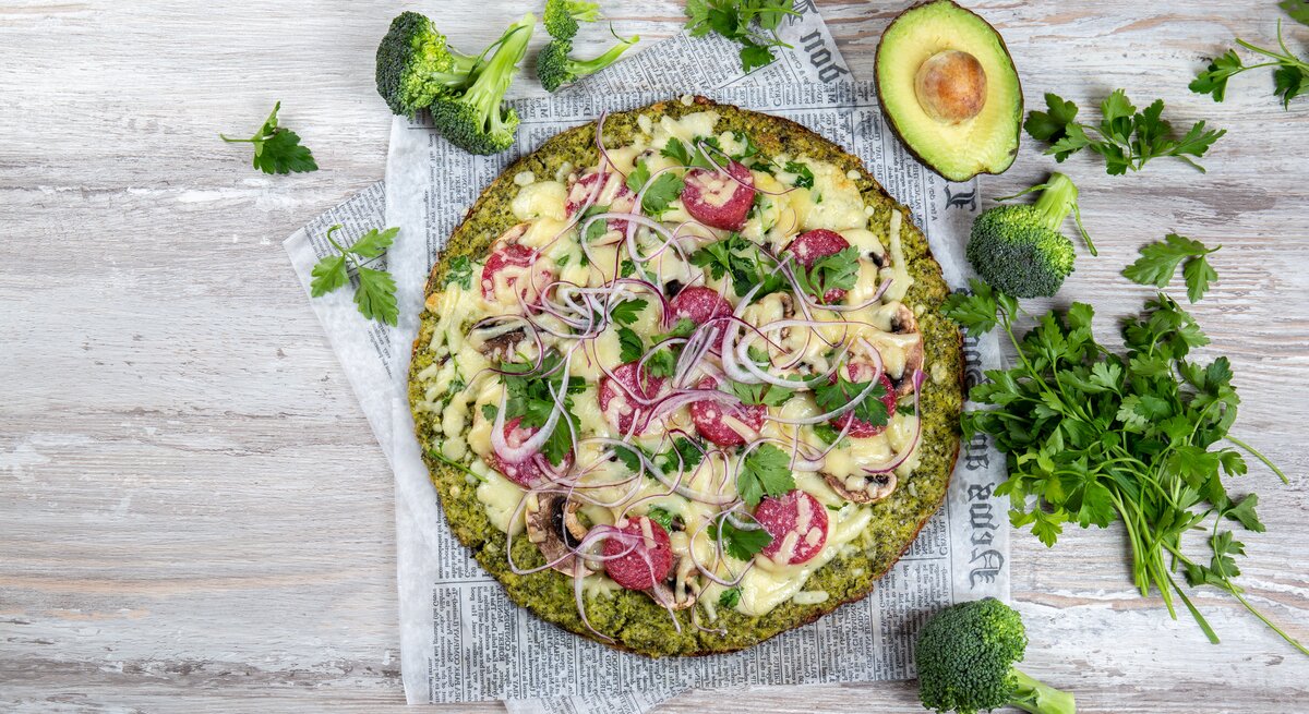 Pizza mit Brokkoliboden, serviert auf Zeitungspapier, belegt mit Salamischeiben, roten Zwiebeln und Käse.