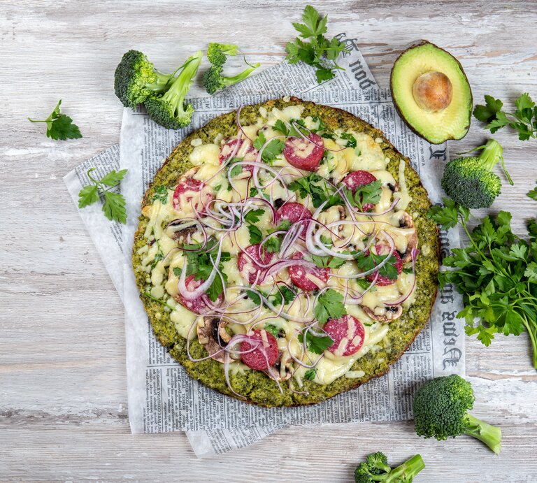 Pizza mit Brokkoliboden, serviert auf Zeitungspapier, belegt mit Salamischeiben, roten Zwiebeln und Käse. | © Getty Images/ LarisaBlinova