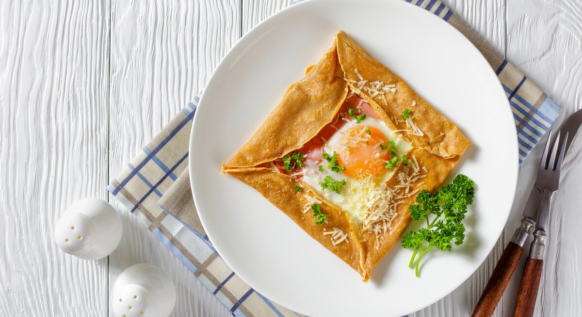 Aus der Vogelperspektive: Ein Teller mit einem Buchweizen-Pfannkuchen mit Schinken, Käse und Spiegelei  steht auf einem Küchentuch. Das Tuch liegt auf einem weißen Holztisch. Daneben stehen zwei Salzstreuer. Auch Messer und Gabel liegen dem dem Teller.