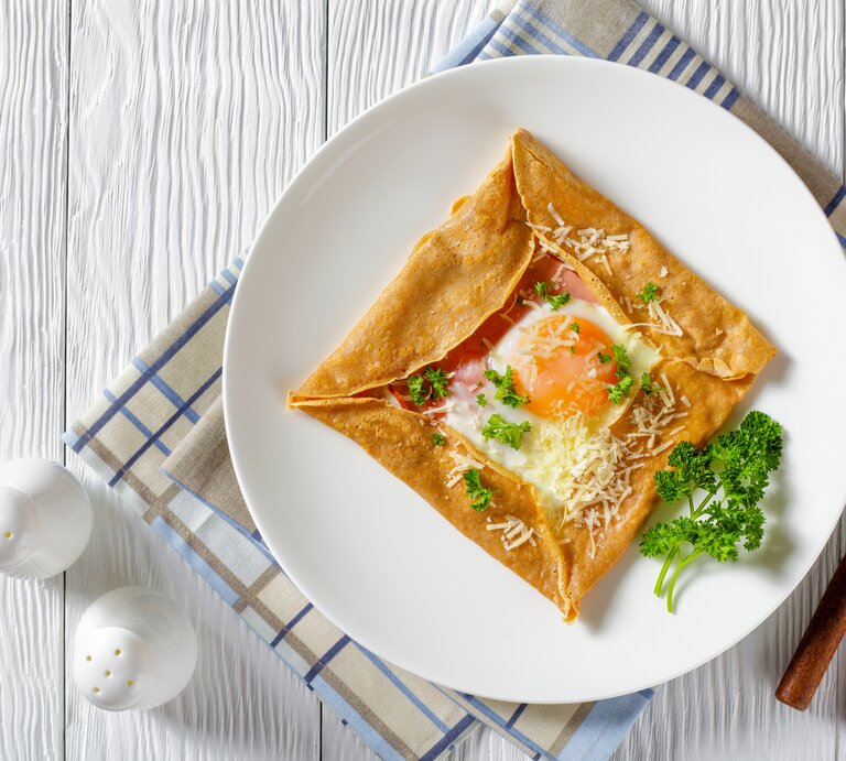 Aus der Vogelperspektive: Ein Teller mit einem Buchweizen-Pfannkuchen mit Schinken, Käse und Spiegelei  steht auf einem Küchentuch. Das Tuch liegt auf einem weißen Holztisch. Daneben stehen zwei Salzstreuer. Auch Messer und Gabel liegen dem dem Teller. | © Getty Images / from_my_point_of_view
