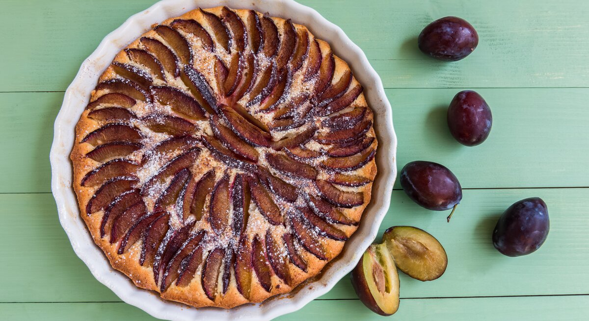 Eine weiße Kuchenform mit dem Pflaumenkuchen auf einem grünen Holztisch direkt von oben fotografiert.