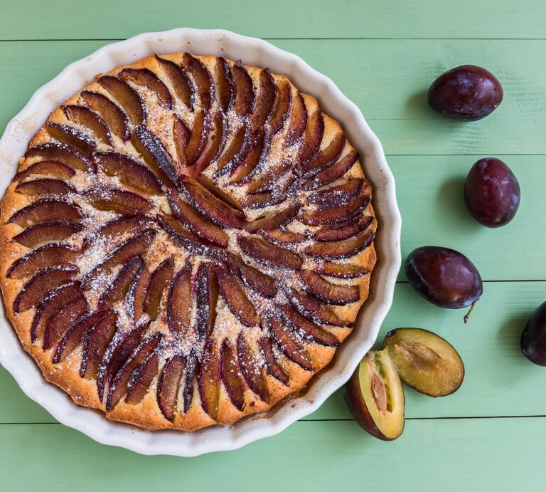 Eine weiße Kuchenform mit dem Pflaumenkuchen auf einem grünen Holztisch direkt von oben fotografiert. | © istock/eugeniek