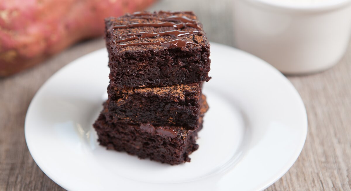 Drei Süßkartoffel-Brownies auf einem weißen Teller in der Nahaufnahme. Alles auf grauer Tischfläche und eine Süßkartoffel unscharf im Hintergrund erkennbar.