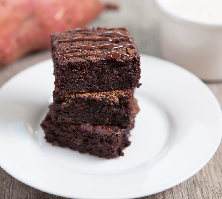 Drei Süßkartoffel-Brownies auf einem weißen Teller in der Nahaufnahme. Alles auf grauer Tischfläche und eine Süßkartoffel unscharf im Hintergrund erkennbar. | © istock/Aiselin82