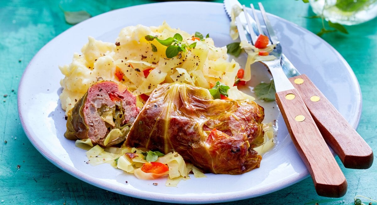 Spitzkohl-Rouladen an Kartoffelpüree auf einem weißen Teller und blauem Untergrund. Alles von schräg oben fotografiert.