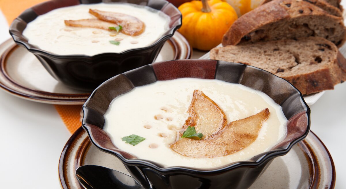 Zwei dunkle Schüsseln mit Pastinaken-Suppe, dekoriert mit Birnenscheiben, Ahornsirup und Petersilie, daneben Brotscheiben und Kürbisse.