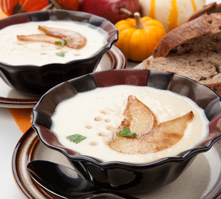 Zwei dunkle Schüsseln mit Pastinaken-Suppe, dekoriert mit Birnenscheiben, Ahornsirup und Petersilie, daneben Brotscheiben und Kürbisse. | © Getty Images/ evgenyb