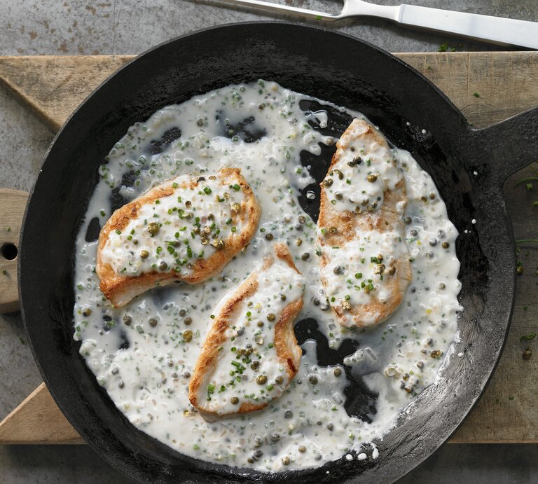 Eine gusseiserne Pfanne mit dem Hähnchen in Rahmsoße auf einem Holzbrett und grauem Tisch direkt von oben fotografiert. | © StockFood / Westermann, Jan-Peter