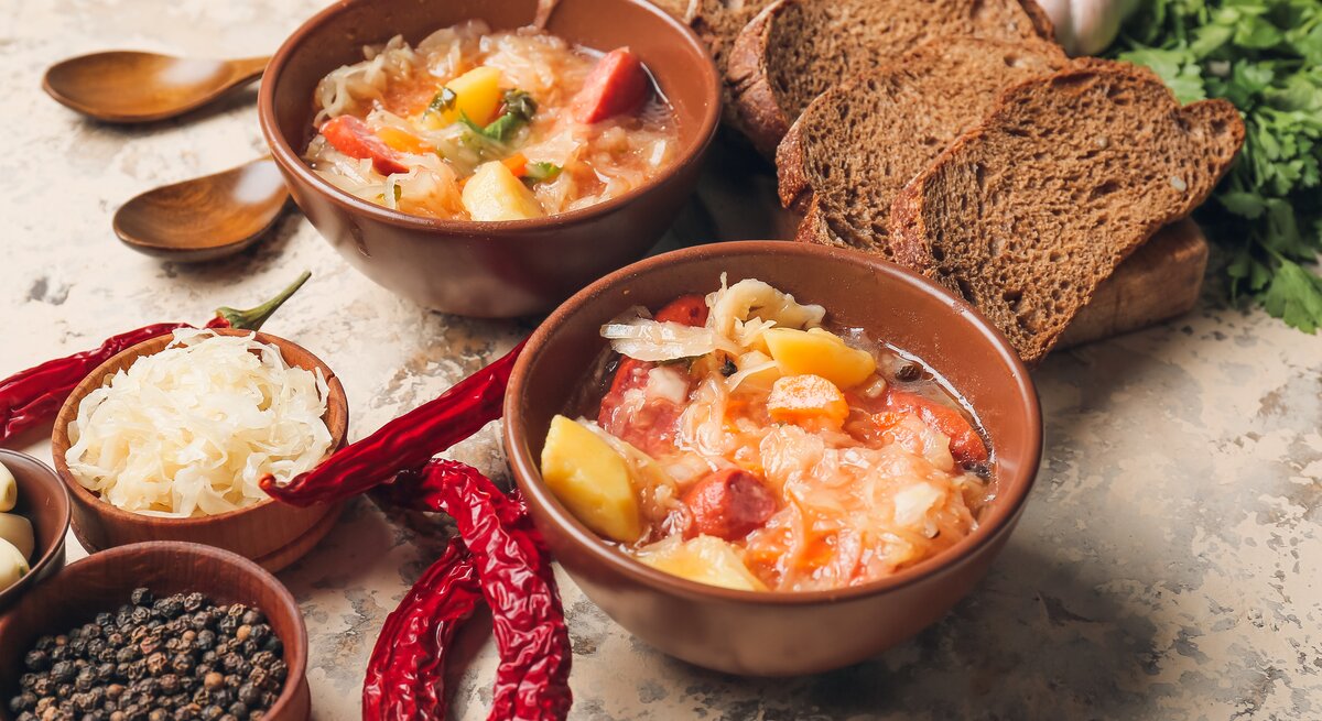 Draufsicht: Auf einem Untergrund aus Mamor stehen zwei Schüsseln mit einer Sauerkraut-Kassler-Suppe. Drum herum liegen Brotscheiben, Holzlöffel, rote Chilischoten, eine Schale Pfefferkörner und frisches Sauerkraut.