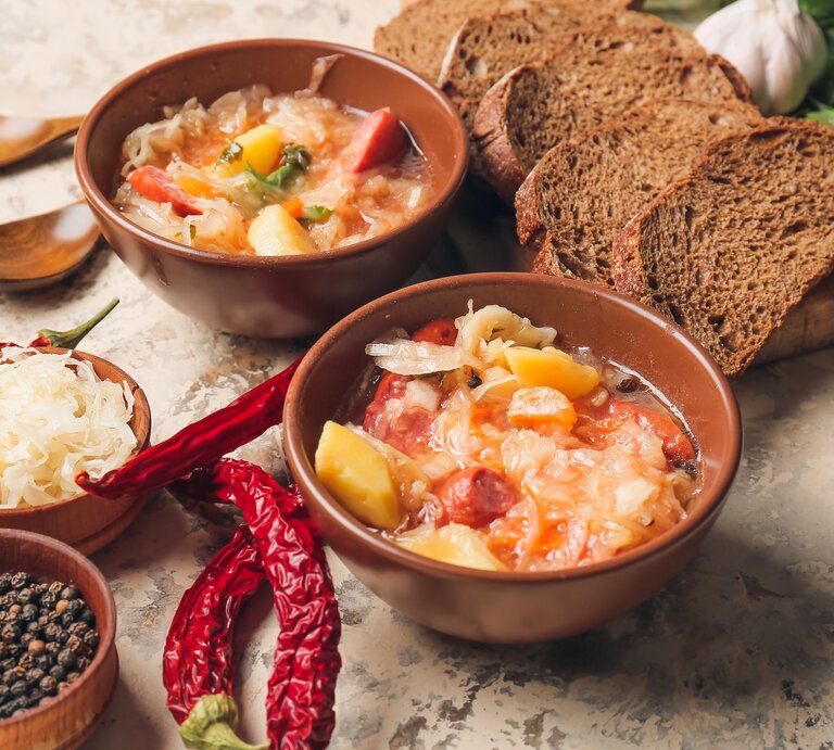 Draufsicht: Auf einem Untergrund aus Mamor stehen zwei Schüsseln mit einer Sauerkraut-Kassler-Suppe. Drum herum liegen Brotscheiben, Holzlöffel, rote Chilischoten, eine Schale Pfefferkörner und frisches Sauerkraut. | © Shutterstock / Pixel-Shot