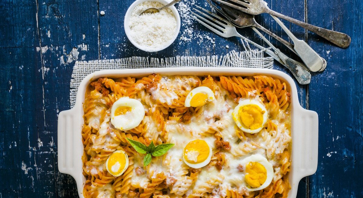 Weiße Form mit Nudelauflauf mit Eiern, daneben Besteck und Parmesan 