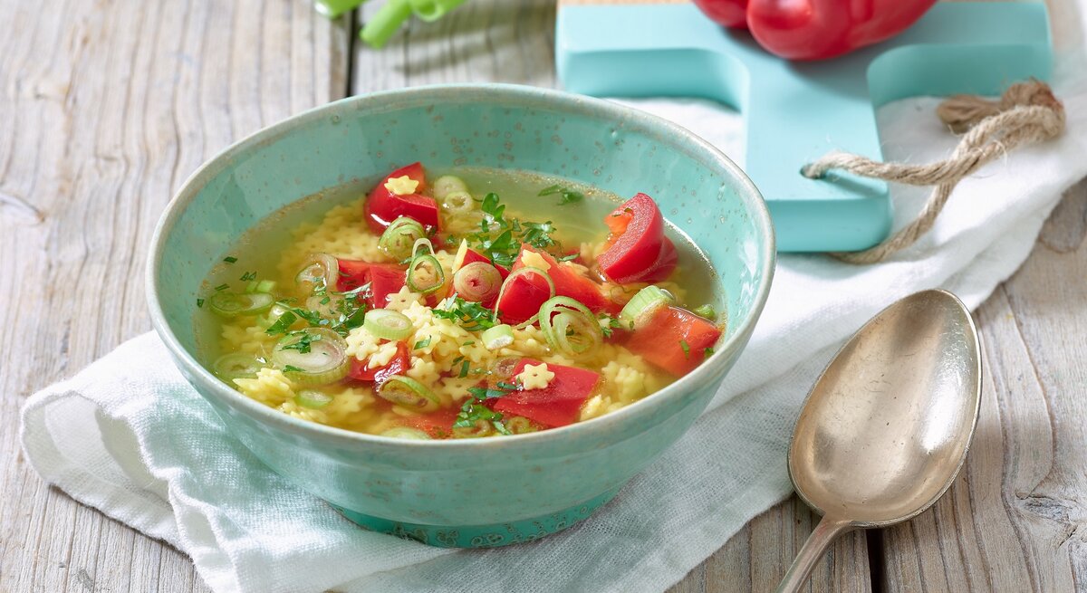 Eine dampfende Schüssel klare Nudelsuppe mit roten Paprikawürfeln, grünen Lauchzwiebelringen und Sternchennudeln.