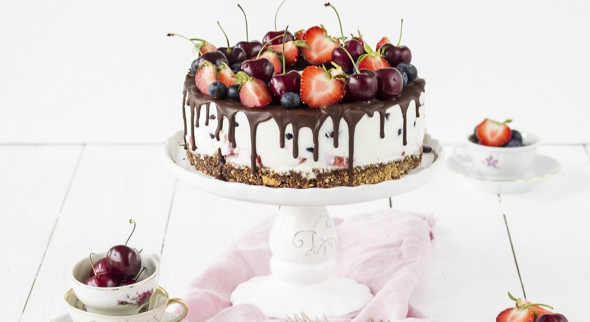Weiße Beerentorte mit Kirschen und Erdbeeren auf einem Tortenständer, mit Schokoladenglasur am Rand. Die Torte steht auf einem hellen Holztisch, umgeben von Vintage-Tassen, Besteck und einem rosa Tuch.