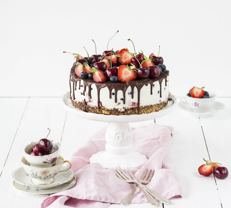 Weiße Beerentorte mit Kirschen und Erdbeeren auf einem Tortenständer, mit Schokoladenglasur am Rand. Die Torte steht auf einem hellen Holztisch, umgeben von Vintage-Tassen, Besteck und einem rosa Tuch. | © StockFood / Friedrichs, Emma