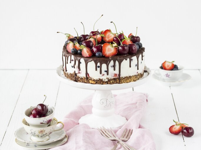 Weiße Beerentorte mit Kirschen und Erdbeeren auf einem Tortenständer, mit Schokoladenglasur am Rand. Die Torte steht auf einem hellen Holztisch, umgeben von Vintage-Tassen, Besteck und einem rosa Tuch. | © StockFood / Friedrichs, Emma