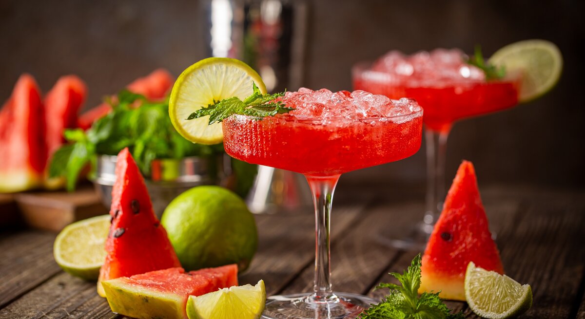 Zwei Gläser Wassermelonen-Daiquiri dekoriert mit Minze und Zitronenscheiben, drumherum frische Minze und Melonenscheiben, dunkler Hintergrund.
