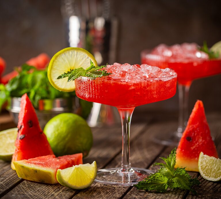 Zwei Gläser Wassermelonen-Daiquiri dekoriert mit Minze und Zitronenscheiben, drumherum frische Minze und Melonenscheiben, dunkler Hintergrund. | © Adobe Stock/ petrrgoskov