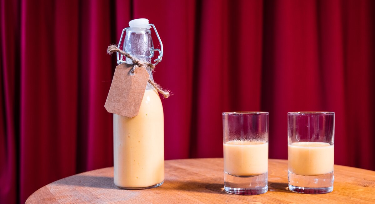 zwei Gläschen Eierlikör, daneben eine kleine Flasche. Das Ganze steht auf einem Holztisch, dahinter ein roter Vorhang.