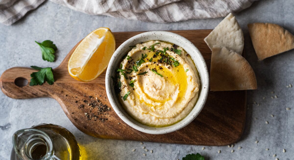 Mangold-Hummus: eine Schale mit Hummus, Olivenöl und Petersilie auf einem Brett, daneben ein Stück Zitrone, einige Scheiben Pitabrot, ein Kännchen mit Öl und ein Handtuch. 