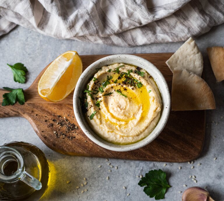 Mangold-Hummus: eine Schale mit Hummus, Olivenöl und Petersilie auf einem Brett, daneben ein Stück Zitrone, einige Scheiben Pitabrot, ein Kännchen mit Öl und ein Handtuch.  | © Getty Images/ Olga Peshkova