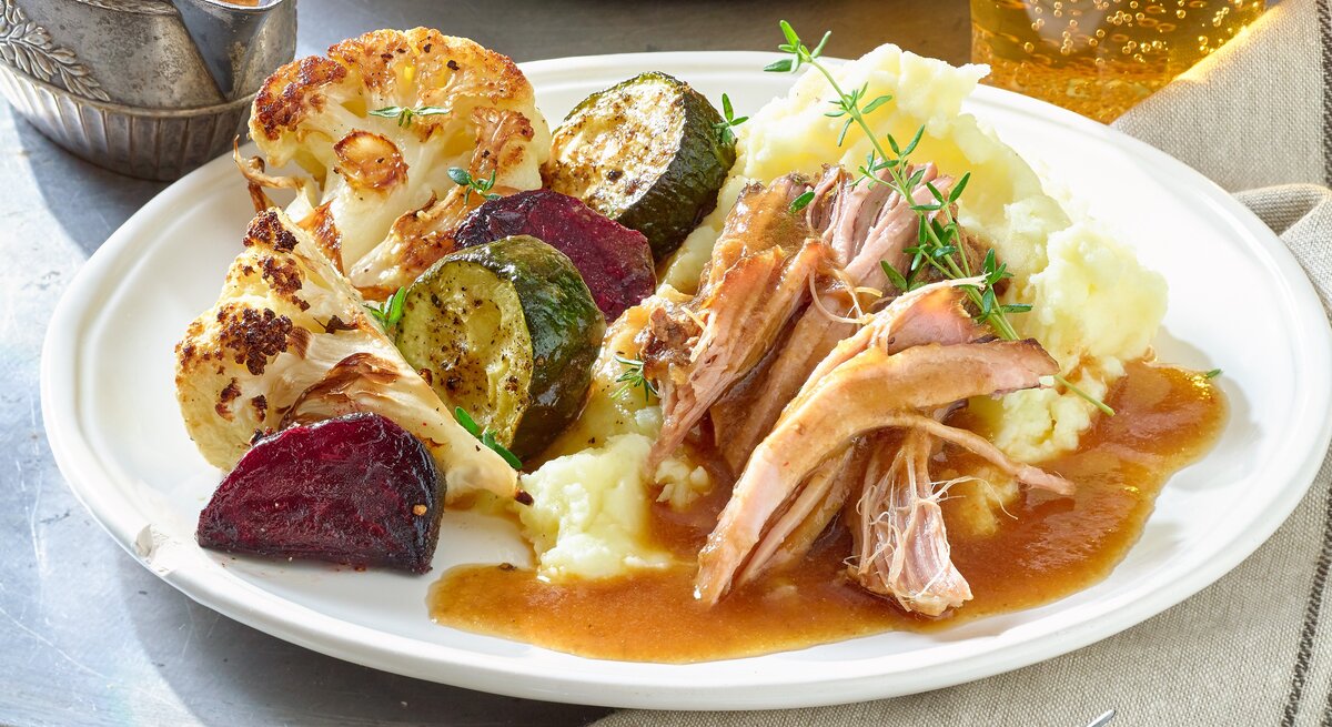 Pulled-Pork-Braten auf einem weißen Teller mit Kartoffelpüree, gebratenem Blumenkohl, Zucchini und roter Bete. Serviert mit Bratensoße. Der Teller steht auf einem Metalltisch neben einem grau-braunen Handtuch.