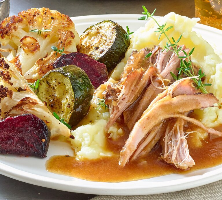 Pulled-Pork-Braten auf einem weißen Teller mit Kartoffelpüree, gebratenem Blumenkohl, Zucchini und roter Bete. Serviert mit Bratensoße. Der Teller steht auf einem Metalltisch neben einem grau-braunen Handtuch. | © Matthias Liebich