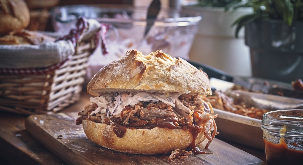 Saftiges Pulled Pork Sandwich mit Krautsalat auf einem rustikalen Holzbrett, im Hintergrund ein geflochtener Brotkorb.