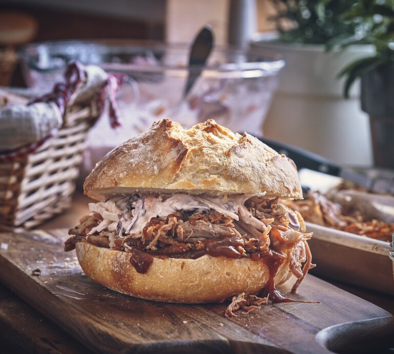 Saftiges Pulled Pork Sandwich mit Krautsalat auf einem rustikalen Holzbrett, im Hintergrund ein geflochtener Brotkorb. | © iStock/GMVozd