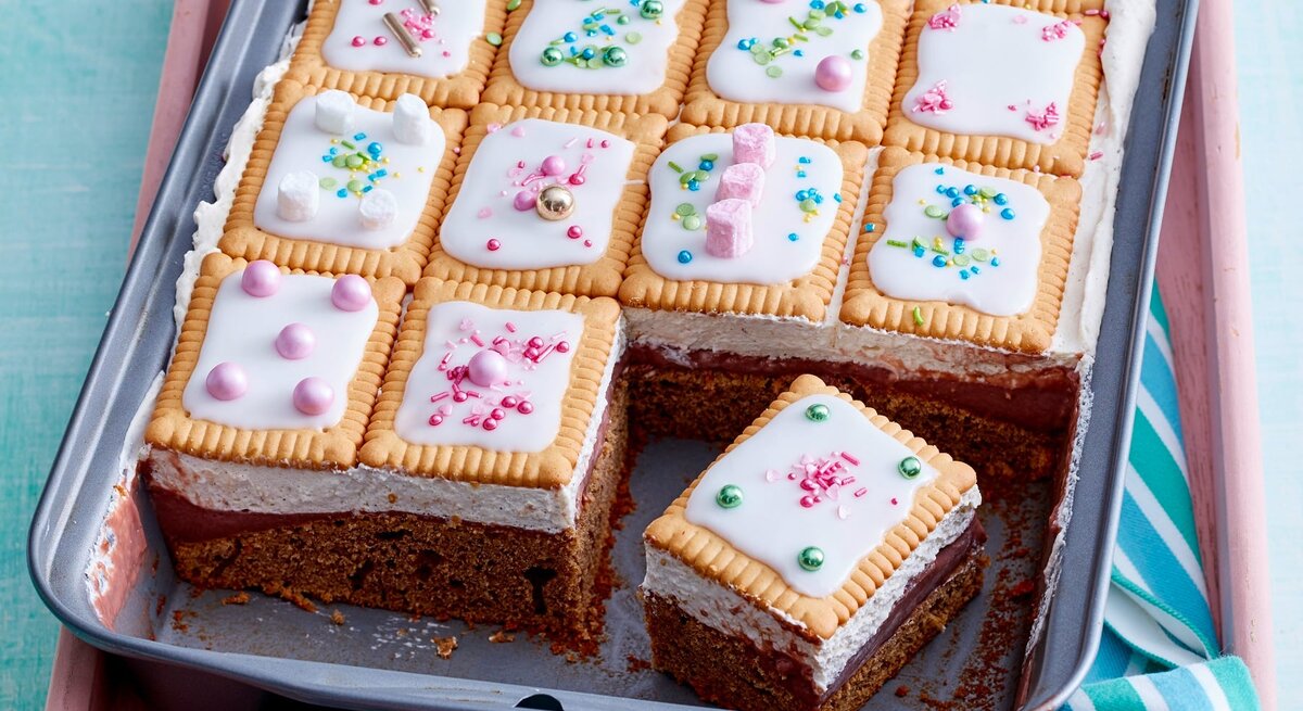 Blechkuchen mit Schokoladenboden, heller Creme und Butterkeksen mit weißem Zuckerguss und bunten Zuckerstreuseln, ein Stück ist herausgeschnitten.