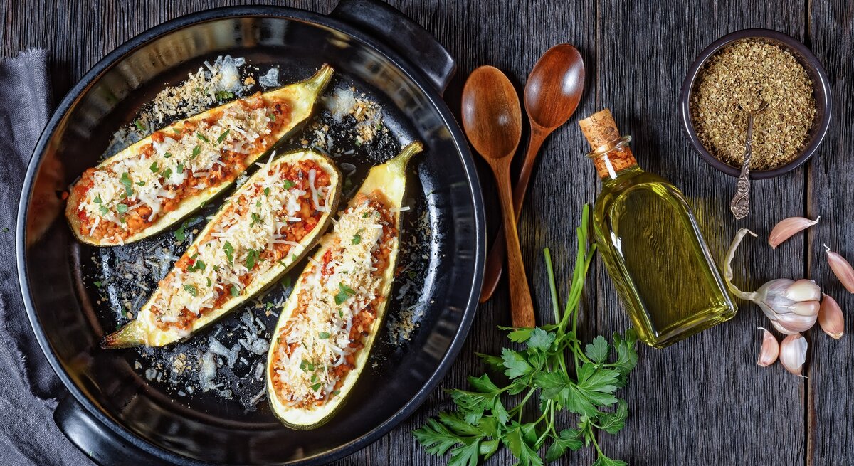 Draufsicht: Gefüllte Zucchini mit Lammhack und Oliven auf einem dunklen Untergrund aus Holz. Daneben liegen zwei Löffel aus Holz, Petersilienzweige, Knoblauchzehen, eine kleine Glasflasche mit Öl und eine schwarze Schale mit einem Gewürz.