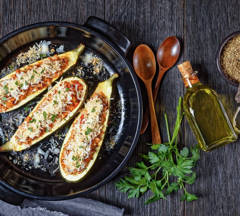 Draufsicht: Gefüllte Zucchini mit Lammhack und Oliven auf einem dunklen Untergrund aus Holz. Daneben liegen zwei Löffel aus Holz, Petersilienzweige, Knoblauchzehen, eine kleine Glasflasche mit Öl und eine schwarze Schale mit einem Gewürz. | © Shutterstock / from my point of view