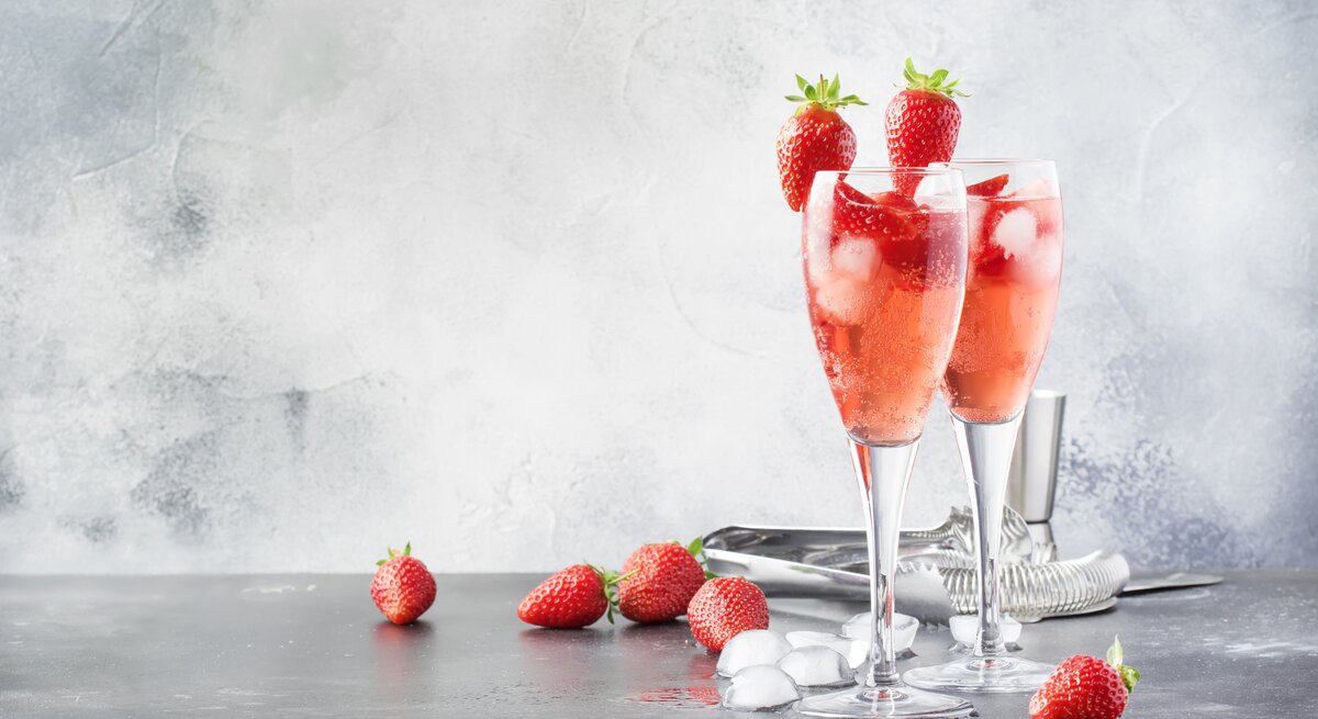 Zwei Gläser mit Rhabarber-Sekt und Erdbeeren stehen auf einem grauen Untergrund. Um die Gläser herum liegen Eiswürfel und Erdbeeren. Hinter den Gläsern liegt eine Zange für die Eiswürfel.