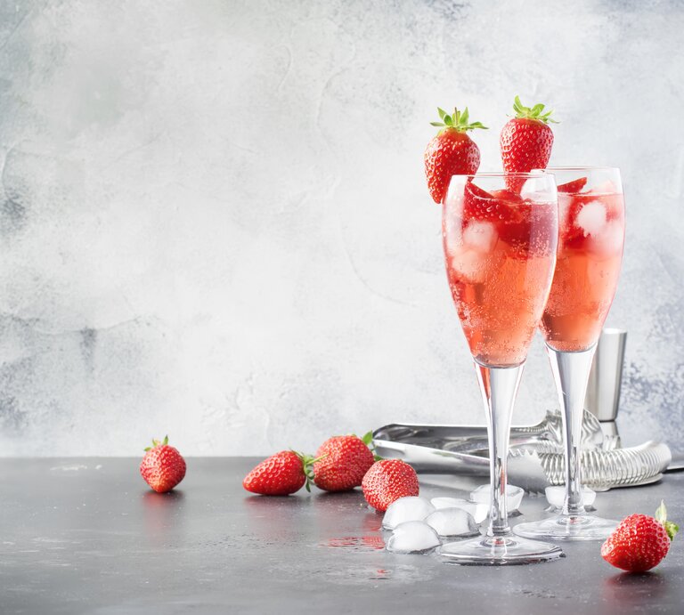 Zwei Gläser mit Rhabarber-Sekt und Erdbeeren stehen auf einem grauen Untergrund. Um die Gläser herum liegen Eiswürfel und Erdbeeren. Hinter den Gläsern liegt eine Zange für die Eiswürfel. | © Getty Images / 5PH / Olga Kochina