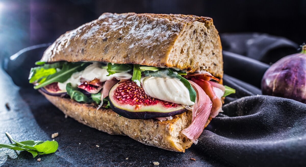 Frontalsicht: Schinken-Mozzarella-Sandwich mit Salatblättern liegt auf einem dunklen Untergrung. Daneben ist ein dunkles Tuch.