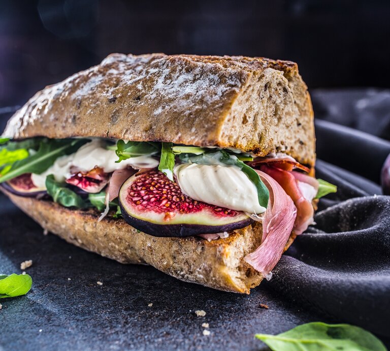 Frontalsicht: Schinken-Mozzarella-Sandwich mit Salatblättern liegt auf einem dunklen Untergrung. Daneben ist ein dunkles Tuch. | © Shutterstock / Marian Weyo