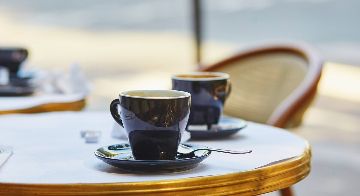 Frontalblick: Zwei French Hot Coffee in Tassen stehen auf einem runden Bistrotisch. An dem Tisch steht ein Stuhl.