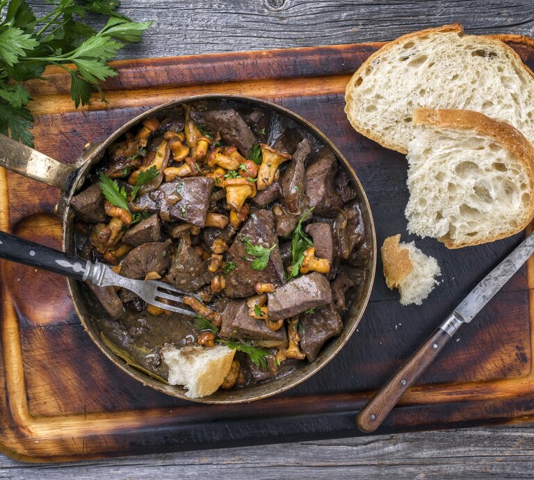 Draufsicht: auf dunklem Untergrund liegt ein Brettchen mit einer rustikalen Pfanne geratene Leber mit Pfifferlingen. | © Shutterstock / hlphoto