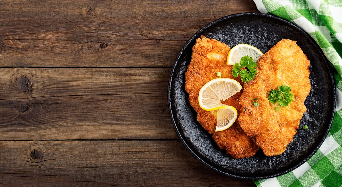 Draufsicht: Auf einem Holztisch steht ein dunkler Teller mit einem Schnitzel mit Brezn-Panade. Die sind mit Peterstilie und Zitronenscheiben dekoriert. Neben dem Teller liegt ein grün-weiß kartiertes Tuch.