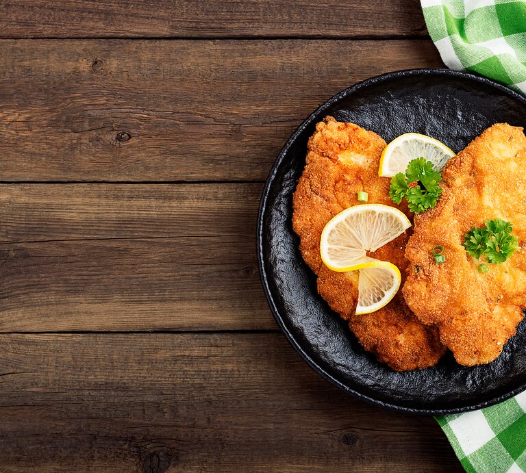 Draufsicht: Auf einem Holztisch steht ein dunkler Teller mit einem Schnitzel mit Brezn-Panade. Die sind mit Peterstilie und Zitronenscheiben dekoriert. Neben dem Teller liegt ein grün-weiß kartiertes Tuch. | © Shutterstock / nelea33