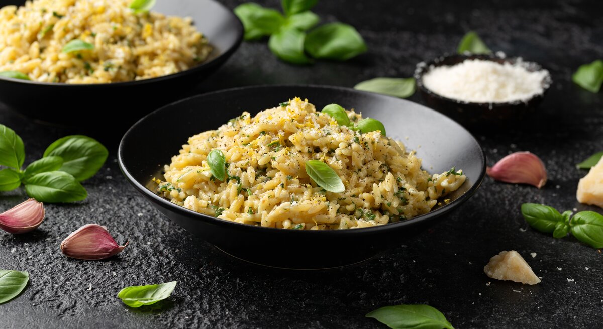 Frontalsicht: Auf einem schwarzen Untergrund stehen zwei dunkle runde Teller. In ihnen ist ein buttriges Zitronen-Orzo mit Parmesan. Drum herum liegen Basilikumblätter und Knoblauchzehen.