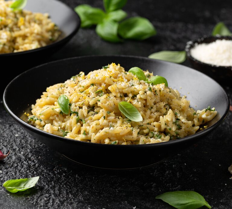 Frontalsicht: Auf einem schwarzen Untergrund stehen zwei dunkle runde Teller. In ihnen ist ein buttriges Zitronen-Orzo mit Parmesan. Drum herum liegen Basilikumblätter und Knoblauchzehen. | © Shutterstock / DronG