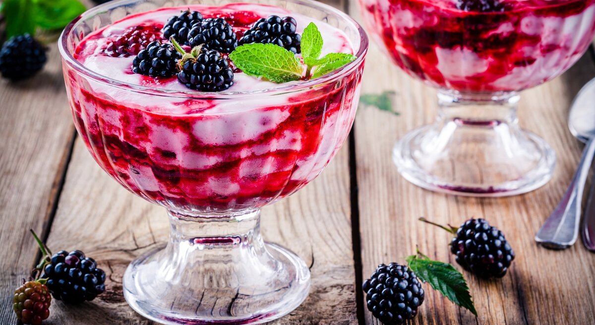 Frontal: Untergrund aus Holz, zwei Dessert-Gläser Brombeer-Vanille-Creme und einige lose Brombeeren sowie ein Dessertlöffel.