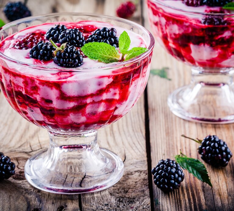 Frontal: Untergrund aus Holz, zwei Dessert-Gläser Brombeer-Vanille-Creme und einige lose Brombeeren sowie ein Dessertlöffel. | © Shutterstock / nblx