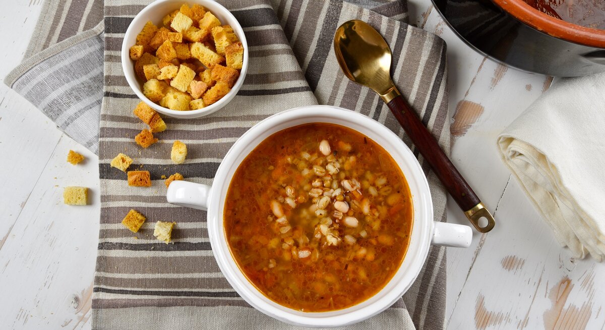 Draufsicht: Eine weiße Schüssel mit einer Kartoffelsuppe mit weißen Bohnen steht auf einem Küchenhandtuch, dass in verschiedenen Brauntönen gestreift ist. Daneben ist eine kleine Schüssel mit Brotwürfeln und einem Löffel. Drum herum stehen eine Schüssel dunkle Brühe sowie ein weißes Tuch.
