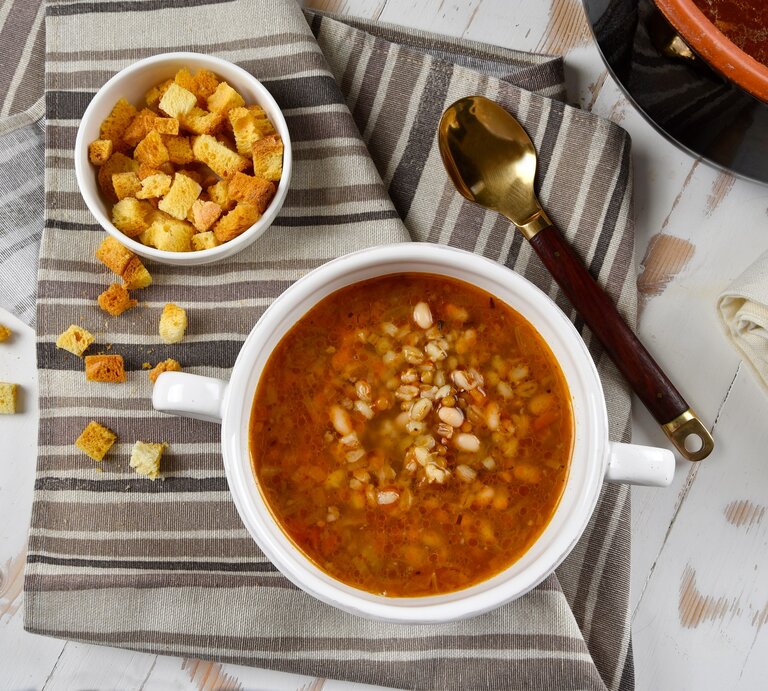 Draufsicht: Eine weiße Schüssel mit einer Kartoffelsuppe mit weißen Bohnen steht auf einem Küchenhandtuch, dass in verschiedenen Brauntönen gestreift ist. Daneben ist eine kleine Schüssel mit Brotwürfeln und einem Löffel. Drum herum stehen eine Schüssel dunkle Brühe sowie ein weißes Tuch. | © Shutterstock / barbajones