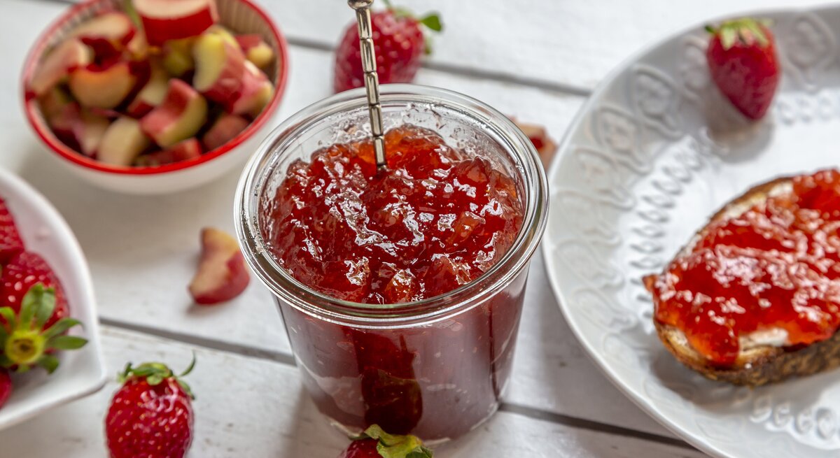 Draufsicht, schräg: Ein Glas Rhabarber-Erdbeer-Gelee steht auf einem weißen Untergrund aus Holz. Daneben liegen Erdbeeren, Rhabarerstücke sowie ein weißer Teller mit einem weißen Teller, auf dem eine Scheibe Brot mit Rhabarber-Erdbeer-Gelee liegt.