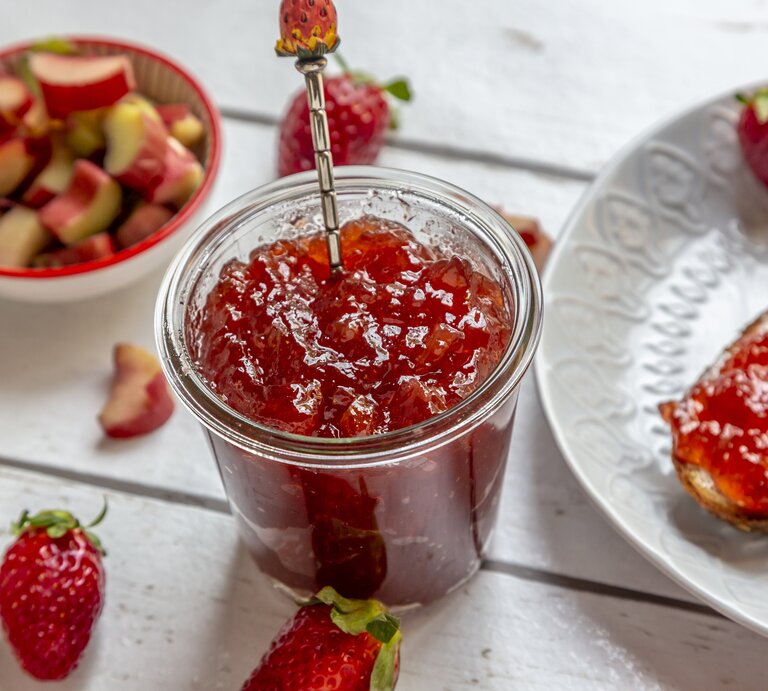 Draufsicht, schräg: Ein Glas Rhabarber-Erdbeer-Gelee steht auf einem weißen Untergrund aus Holz. Daneben liegen Erdbeeren, Rhabarerstücke sowie ein weißer Teller mit einem weißen Teller, auf dem eine Scheibe Brot mit Rhabarber-Erdbeer-Gelee liegt. | © Getty Images / 	Westend61