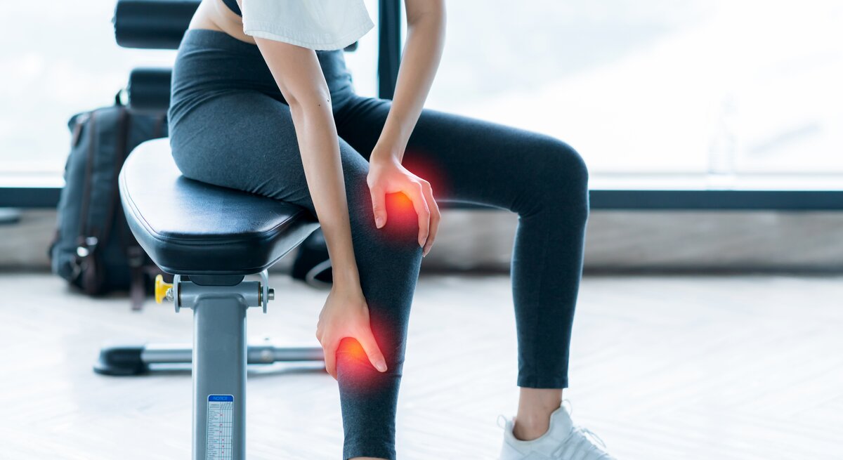 Eine Frau in sportlicher Kleidung sitzt auf einem Trainingsgerät und hält ihr Knie, welches digital rot hervorgehoben ist, um Schmerzen zu symbolisieren. Sie trägt ein weißes Handtuch über der Schulter.