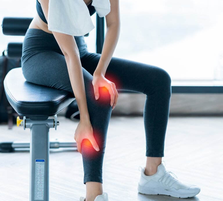 Eine Frau in sportlicher Kleidung sitzt auf einem Trainingsgerät und hält ihr Knie, welches digital rot hervorgehoben ist, um Schmerzen zu symbolisieren. Sie trägt ein weißes Handtuch über der Schulter. | © Getty Images/whyframestudio